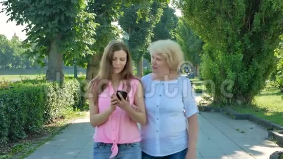 快乐的老年妇女和年轻妇女步行公园车道使用智能手机成年母亲和女儿在公园散步视频的预览图