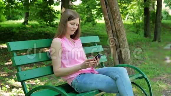 年轻女子坐在公园的长凳上用智能手机在露天使用智能手机的漂亮女孩视频的预览图