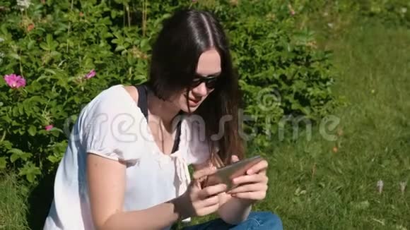 在晴天女人坐在公园里玩手机游戏视频的预览图
