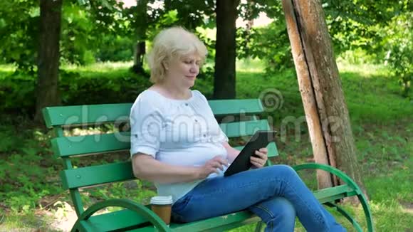 快乐的老妇人坐在公园的长凳上用平板电脑喝咖啡使用平板电脑的高级女性视频的预览图