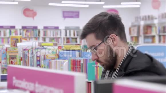年轻的黑发男子戴着眼镜在书店的书架上看书视频的预览图