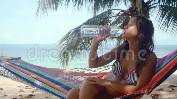 长荣漂亮的女人躺在吊床上喝着海滩上瓶子里的水慢动作1920x1080视频的预览图