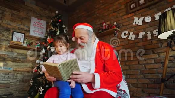 圣诞老人坐在扶手椅上在装饰的节日房间里为热情的小女孩读着童话书视频的预览图