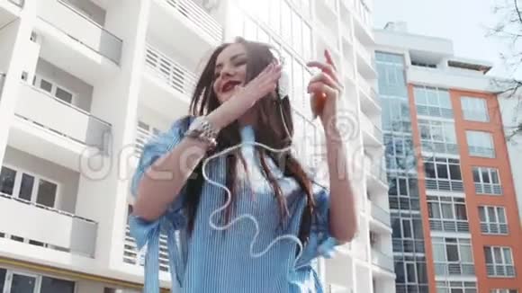 迷人的年轻女子穿着时尚的外观和白色耳机在城市里自由地听音乐和跳舞自然视频的预览图