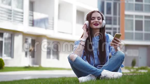 年轻女子穿着休闲牛仔裤穿着优雅的蓝色衬衫穿着白色运动鞋坐在绿草上用她的手机打电话视频的预览图
