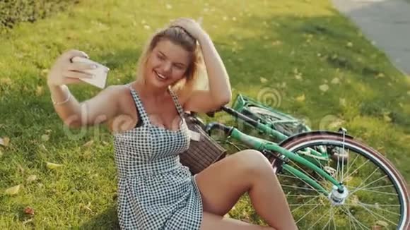 美丽的金发女郎灿烂的微笑坐在城市公园的绿色草坪上抚摸着她的头发拿着她的头发视频的预览图
