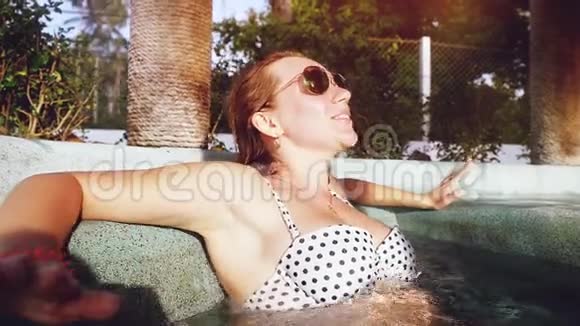年轻漂亮的快乐女人戴着太阳镜跳舞在按摩浴缸里唱歌在室外游泳池享受她的生活视频的预览图
