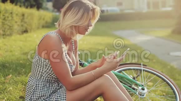 漂亮的金发女郎穿着一件时髦的白色连衣裙坐在城市公园的草坪上旁边是绿色的复古风格视频的预览图