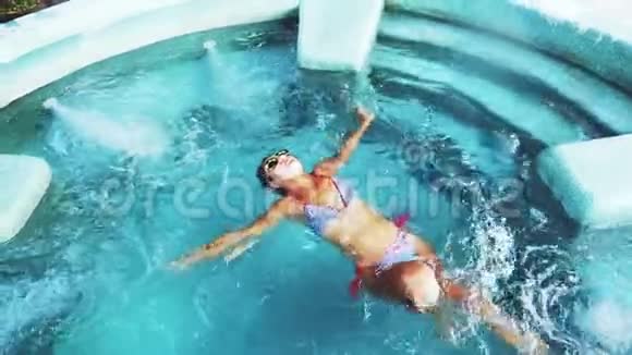 美丽的棕褐色年轻女子穿着比基尼和太阳镜在清澈的游泳池游泳视频的预览图