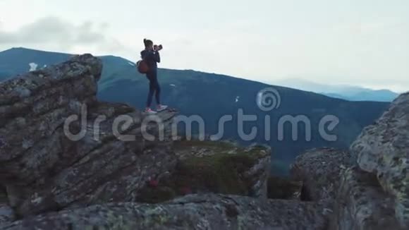 年轻时尚的女摄影师带着背包站在高高的岩石窗台上拿着相机俯瞰绿色的景色视频的预览图