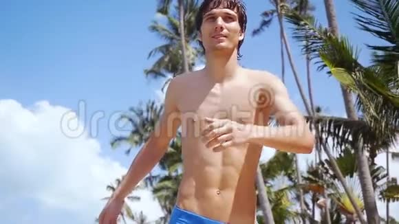 在阳光明媚的日子里年轻英俊的快乐男子在海滩上奔跑在蓝天上的棕榈树上奔跑视频的预览图