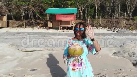 无人驾驶飞机看到年轻漂亮的黑发女人戴着太阳镜手里拿着椰子在海底的海滩上挥舞着手视频的预览图
