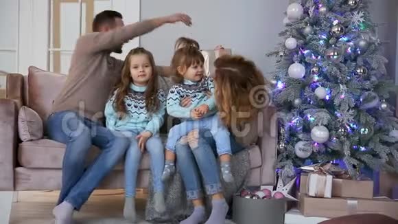 圣诞节快乐的一家人妈妈爸爸和两个小女孩坐在家中的沙发上靠近圣诞树打开视频的预览图