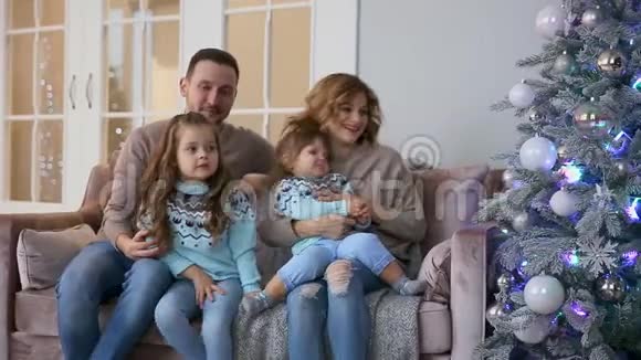 圣诞节快乐的一家人妈妈爸爸和两个小女孩坐在家中的沙发上靠近圣诞树打开视频的预览图
