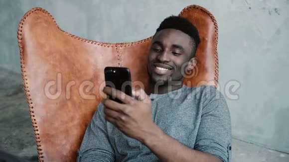 非洲男人坐在椅子上使用智能手机的肖像男性正在上网微笑视频的预览图