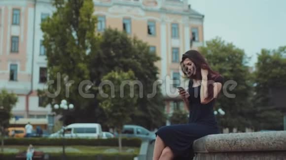 美丽的年轻黑发女人穿着一件黑色的长裙坐在喷泉上积极地用手机聊天视频的预览图