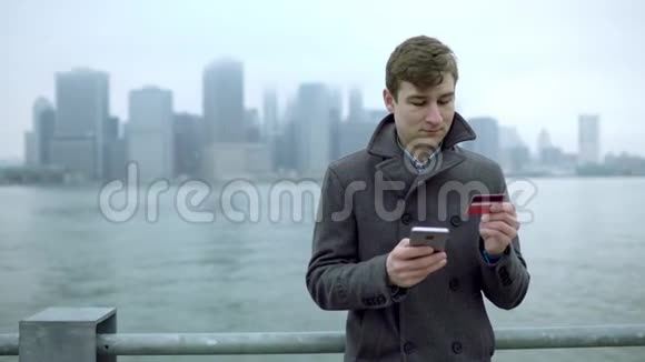 男子通过使用智能手机和信用卡在线购物同时站在哈德逊河附近观看曼哈顿视频的预览图