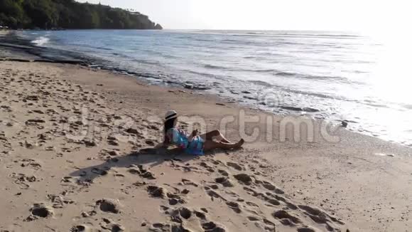 无人驾驶飞机观看戴墨镜的年轻美女躺在热带天堂沙滩上使用手机视频的预览图