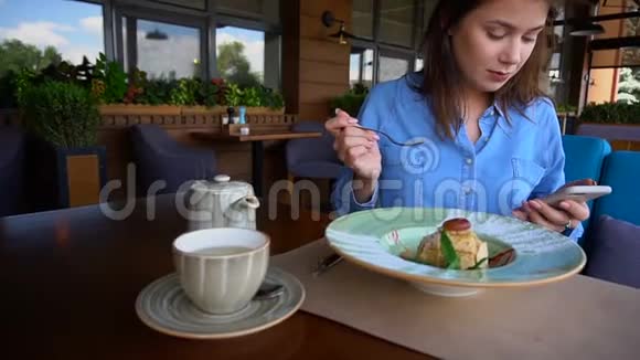 快乐的女孩通过智能手机在咖啡馆慢动作浏览和吃甜点视频的预览图