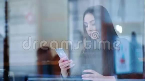 美丽的女孩在一家舒适的A咖啡馆里温暖着一杯热咖啡坐在窗边用她的手机现代生活方式视频的预览图