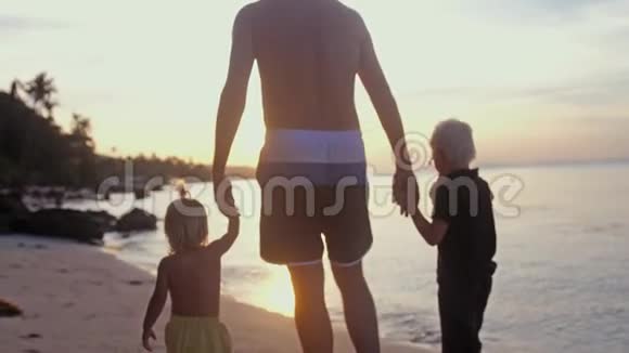 爸爸在美丽夕阳下牵着孩子们的手在热带海滩上散步旅游度假概念慢动作视频的预览图