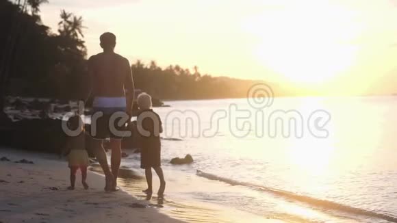父亲和孩子们在美丽的夕阳下在海滩上散步爸爸牵着孩子们手慢动作旅行假期视频的预览图