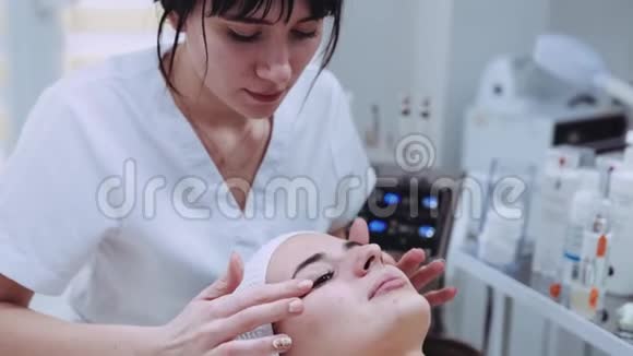 近距离观看一位年轻的专业美容师为一位美丽的女性患者做一个面部整形按摩视频的预览图
