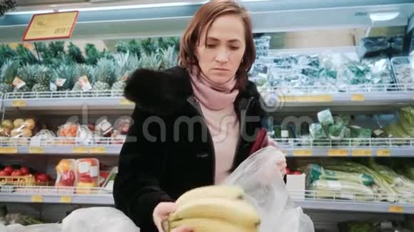 超市里的女人选择香蕉一个穿黑色皮毛大衣的女孩正在商店买香蕉视频的预览图