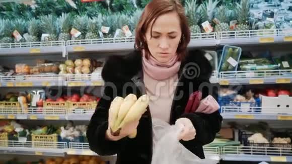 超市里的女人选择香蕉一个穿黑色皮毛大衣的女孩正在商店买香蕉视频的预览图
