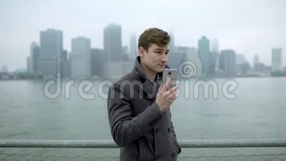 在哈德逊河附近的一个年轻人正在打电话通过他的智能手机交谈可以看到曼哈顿岛视频的预览图