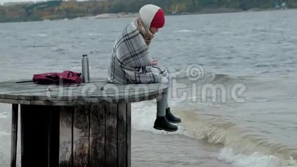 一个穿暖和衣服的年轻女子坐在海边坐在木盘管上喝着热水瓶里的热茶视频的预览图