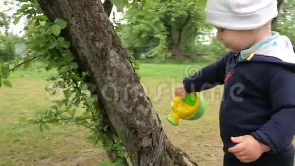 一个可爱的男孩从一个小水罐里倒出一棵大树一个穿着蓝色自行车和白色帽子的孩子在父母花园里视频的预览图