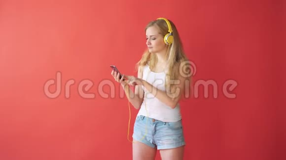 年轻漂亮的白种人女人拿着手机用红色背景的耳机听音乐视频的预览图