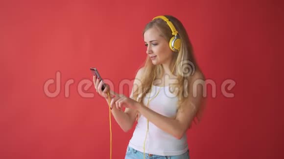 年轻漂亮的白种人女人拿着手机用红色背景的耳机听音乐视频的预览图
