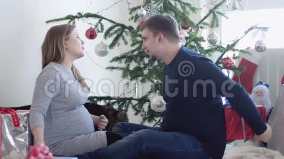 幸福的年轻漂亮夫妇坐在漂亮的圣诞树旁丈夫给怀孕的妻子一个礼品盒视频的预览图