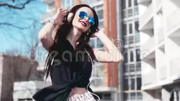 戴着太阳镜的年轻时髦女孩听着耳机里的音乐高兴地随着节奏从一边到另一边跳舞视频的预览图