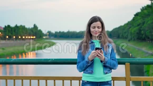 穿着棉质夹克的漂亮女孩站在河边的桥上用智能手机视频的预览图