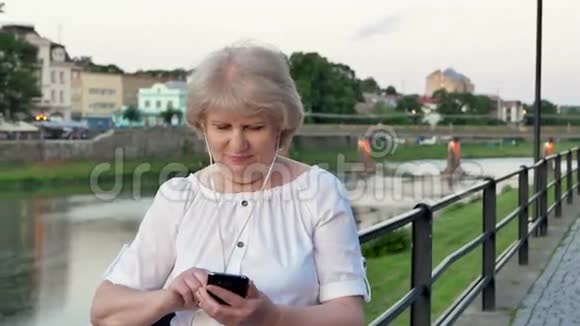傍晚时分在海滨老城区河边和桥边使用带有耳机的智能手机的老年妇女视频的预览图