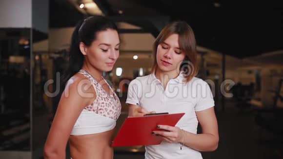 女私人教练在健身房和一个女客户交谈用平板电脑做笔记视频的预览图