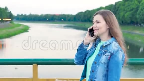 穿着棉袄穿过河上的桥用智能手机说话的漂亮女孩视频的预览图