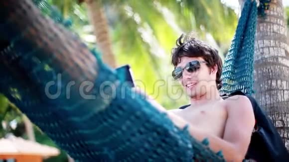 年轻英俊的快乐男人躺在吊床上在美丽的异国海滩上使用智能手机假期概念技术视频的预览图