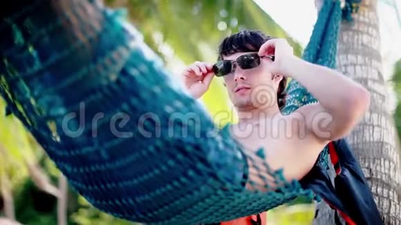 年轻的帅哥戴着太阳镜在美丽的热带海滩上的吊床上放松假期概念1920x1080视频的预览图