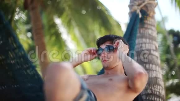 年轻的帅哥戴着太阳镜在美丽的热带海滩上的吊床上放松假期概念1920x1080视频的预览图