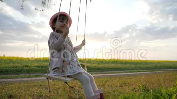 可爱的小女孩肖像坐在大自然的秋千上夕阳的光背景快乐的小孩子玩得很开心视频的预览图
