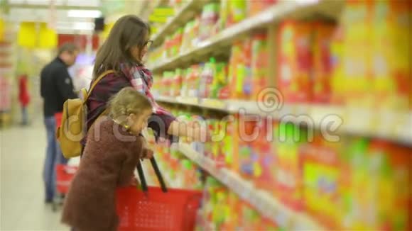 年轻的母亲和小女儿在超市购物妈妈和孩子把产品放在商店的红篮子上视频的预览图