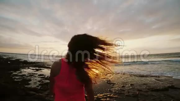 年轻女子站在石质海岸享受日落翅膀吹她的头发晚上海滩上的女孩视频的预览图