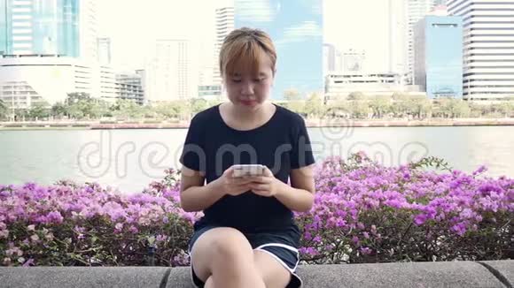 美丽的亚洲年轻女子坐在公园的长凳上用智能手机聊天阅读和发短信视频的预览图