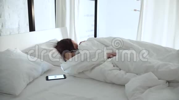 闹钟在电话里疲惫的女人在卧室的床上醒来视频的预览图