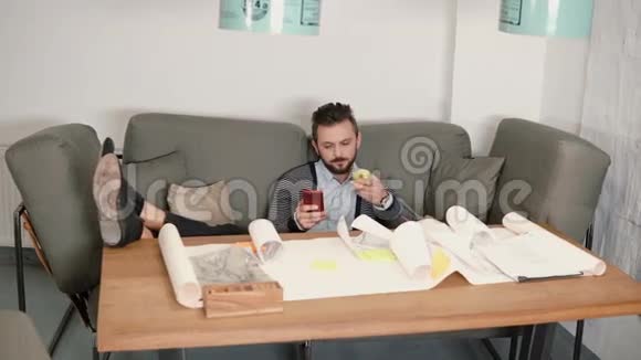 年轻人正在休息避免坐在沙发上工作使用智能手机吃苹果把脚放在桌子上视频的预览图