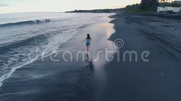 美丽的年轻女性在巴厘岛日落时带着黑色的沙滩和海浪沿着海滩奔跑飞机视频4K视频的预览图
